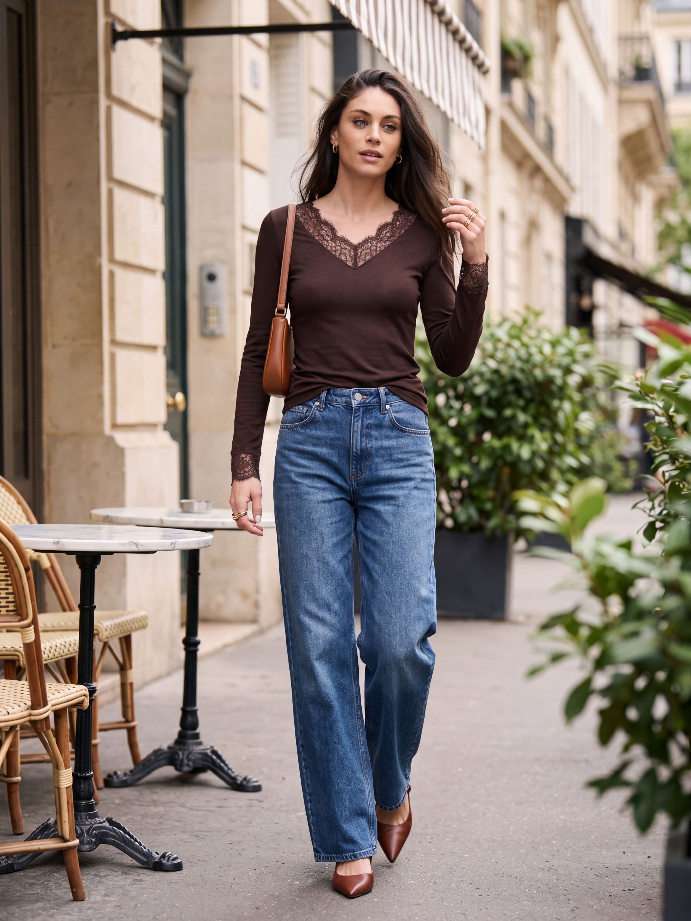 Donna che cammina in città con maglia intima marrone a manica lunga con scollo a V in pizzo, jeans e borsa in pelle