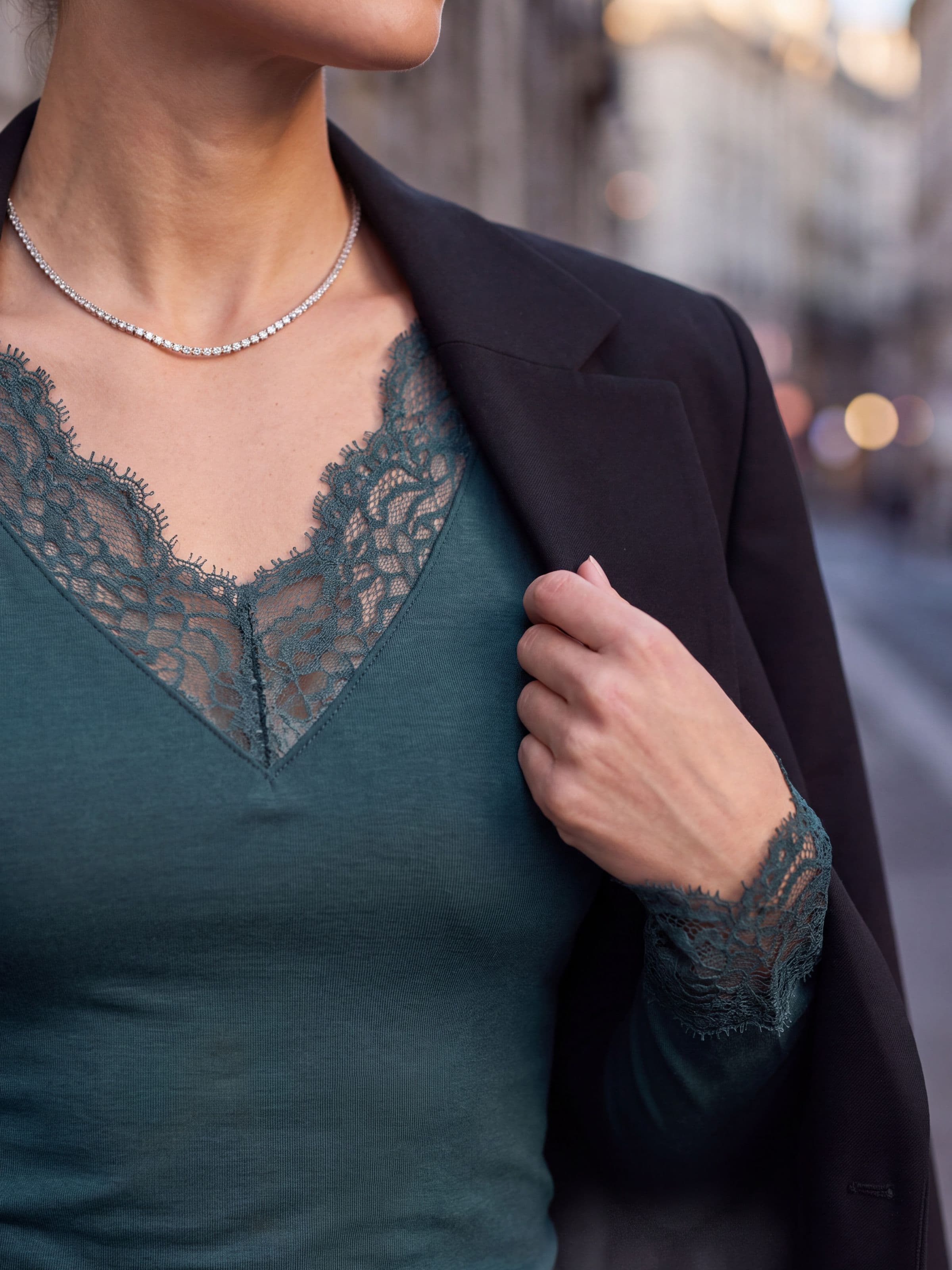 Dettaglio di maglia intima verde bottiglia con scollo a V in pizzo e polsino in pizzo, indossata sotto un blazer nero con una collana