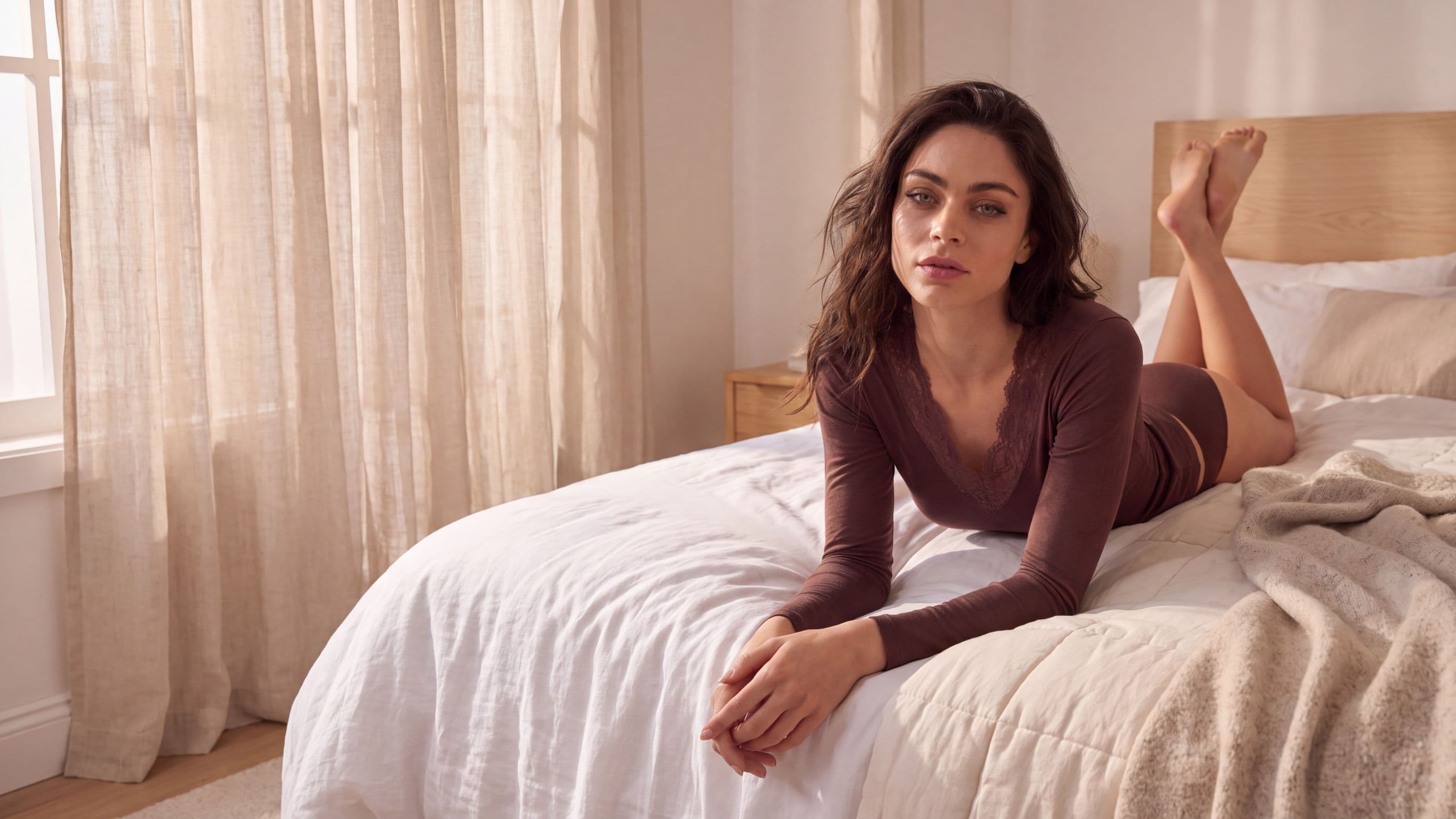Donna sdraiata su un letto in una camera luminosa con maglia intima color prugna con scollo a V in pizzo