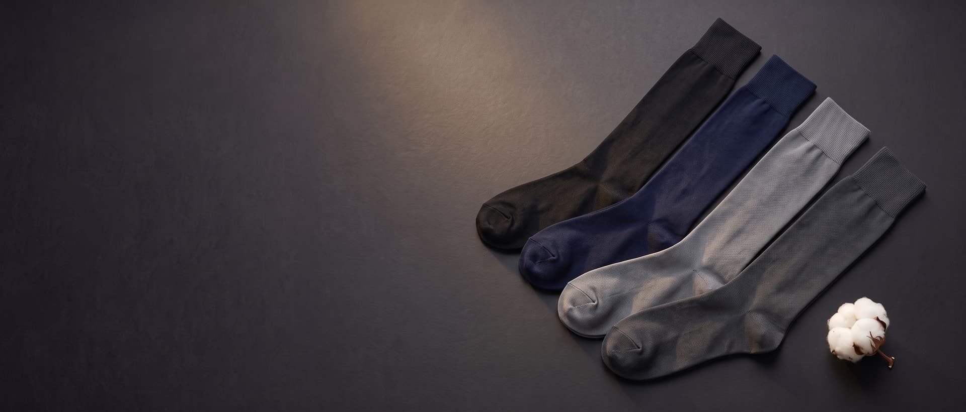 Quattro paia di calze lunghe in Filo di Scozia stese per intero, in nero, blu navy, grigio e antracite, accanto a un fiore di cotone