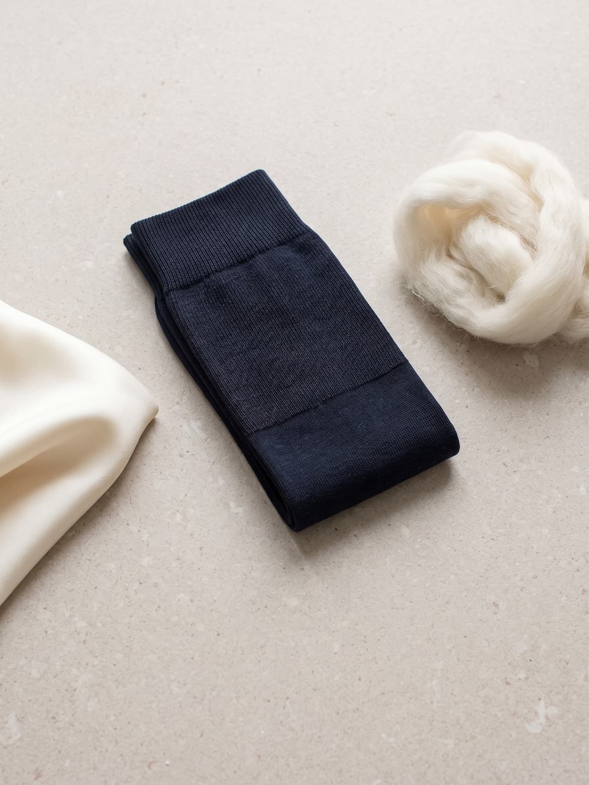 Calza lunga blu navy in lana e seta accanto a un ciuffo di lana Merino e a un tessuto di seta avorio