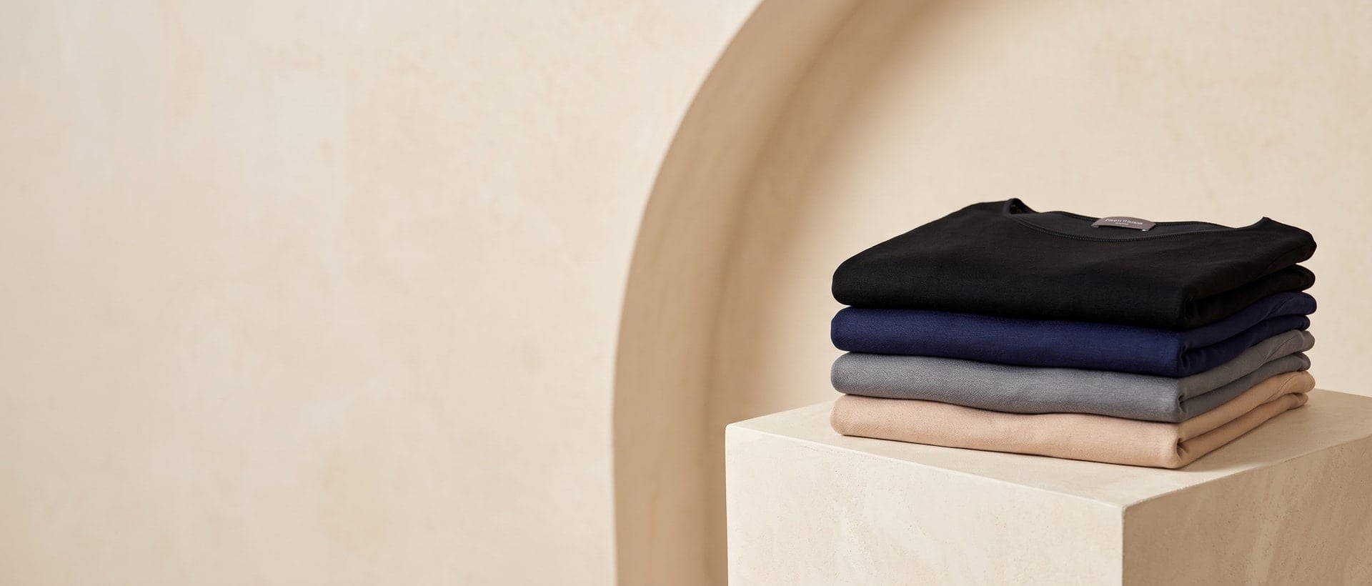 Maglie piegate in nero, blu navy, grigio e beige su un plinto geometrico color panna